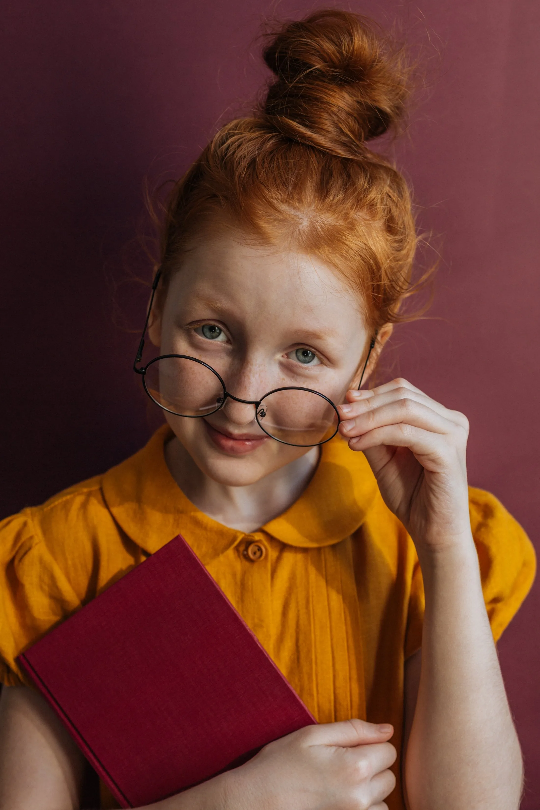 Red-haired girl in glasses and yellow dress holding a red book against a maroon background.