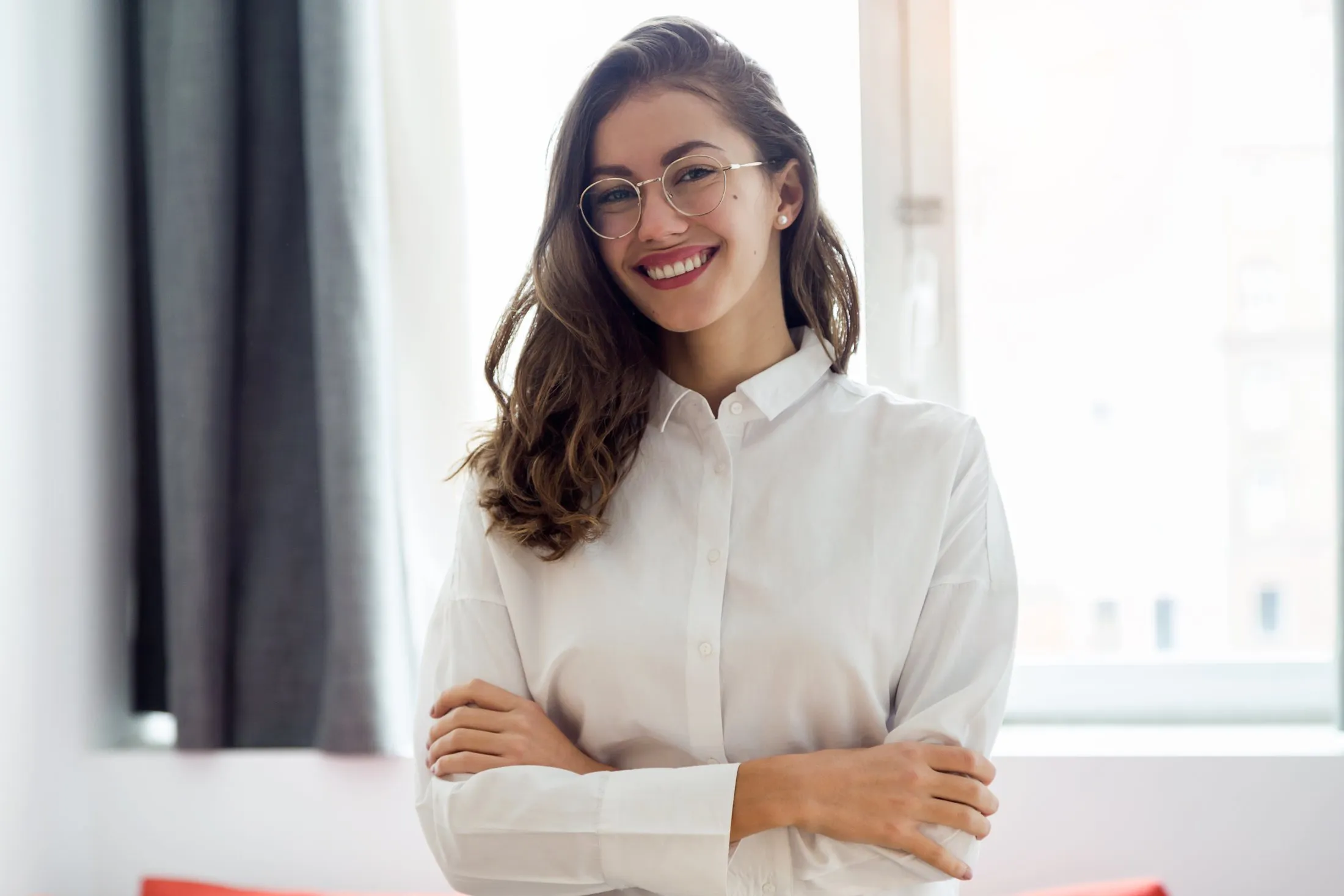 Smiling woman with glasses and long brown hair wearing a white button-up shirt, arms crossed.