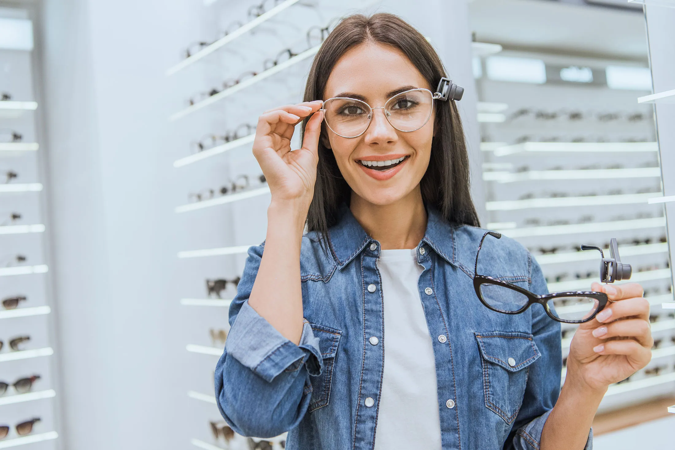 Smiling woman wearing clear glasses holds black-frame glasses inside an eyewear store.