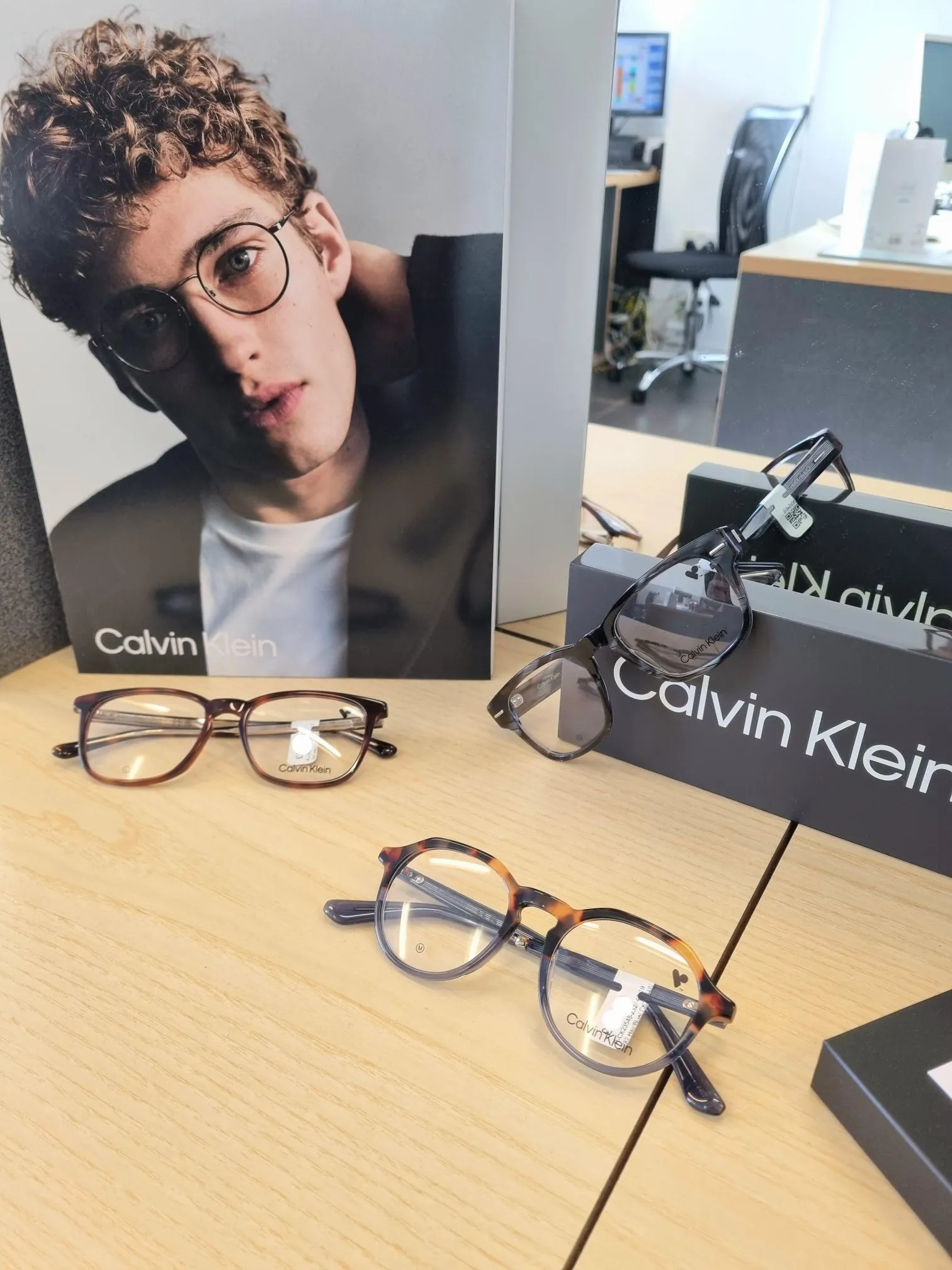 Display of three pairs of Calvin Klein eyeglasses on a wooden table with a Calvin Klein poster in the background.