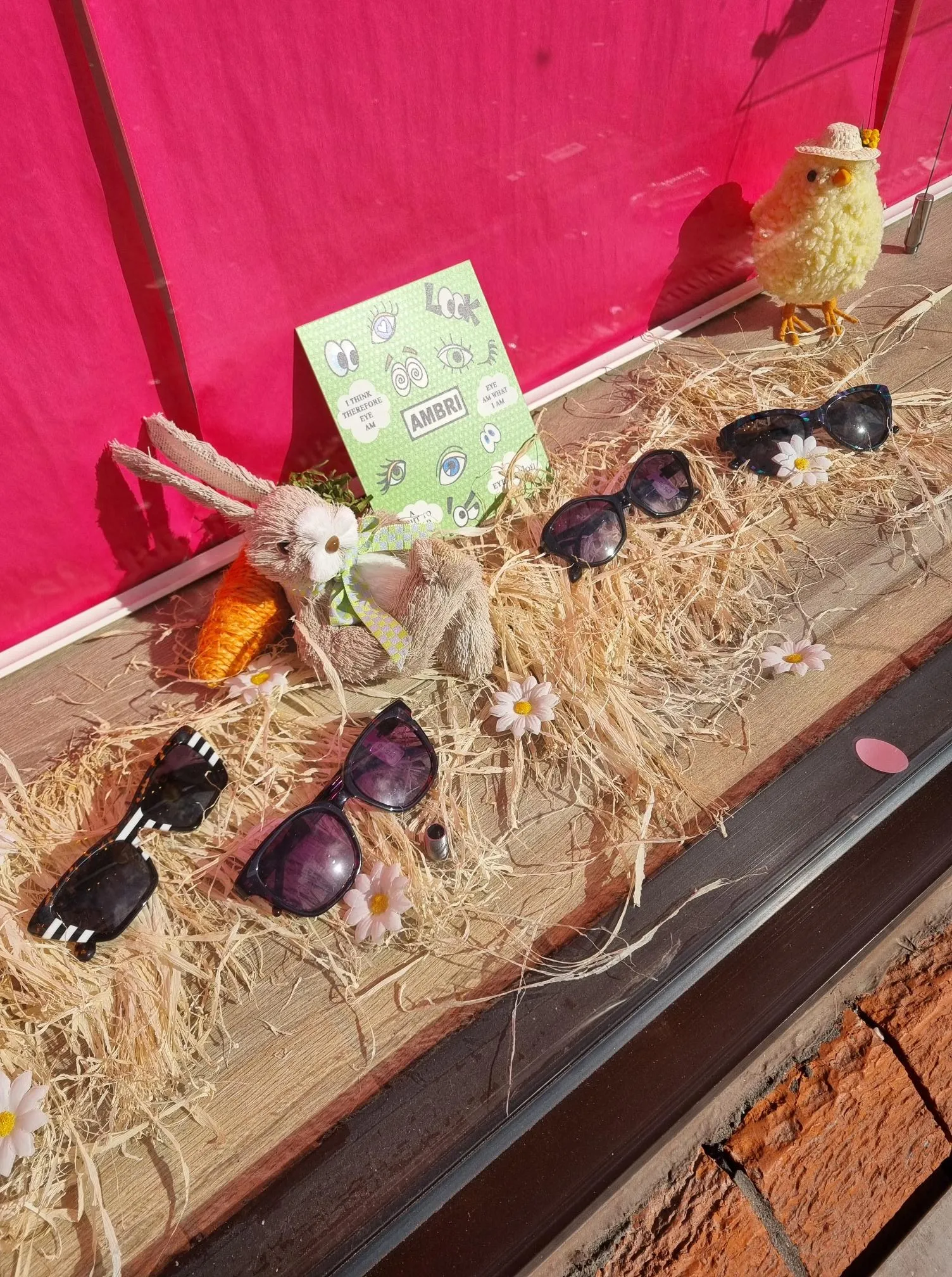 Store display with four sunglasses, a bunny with carrot, a chick figurine, daisies, and a green sign on straw.