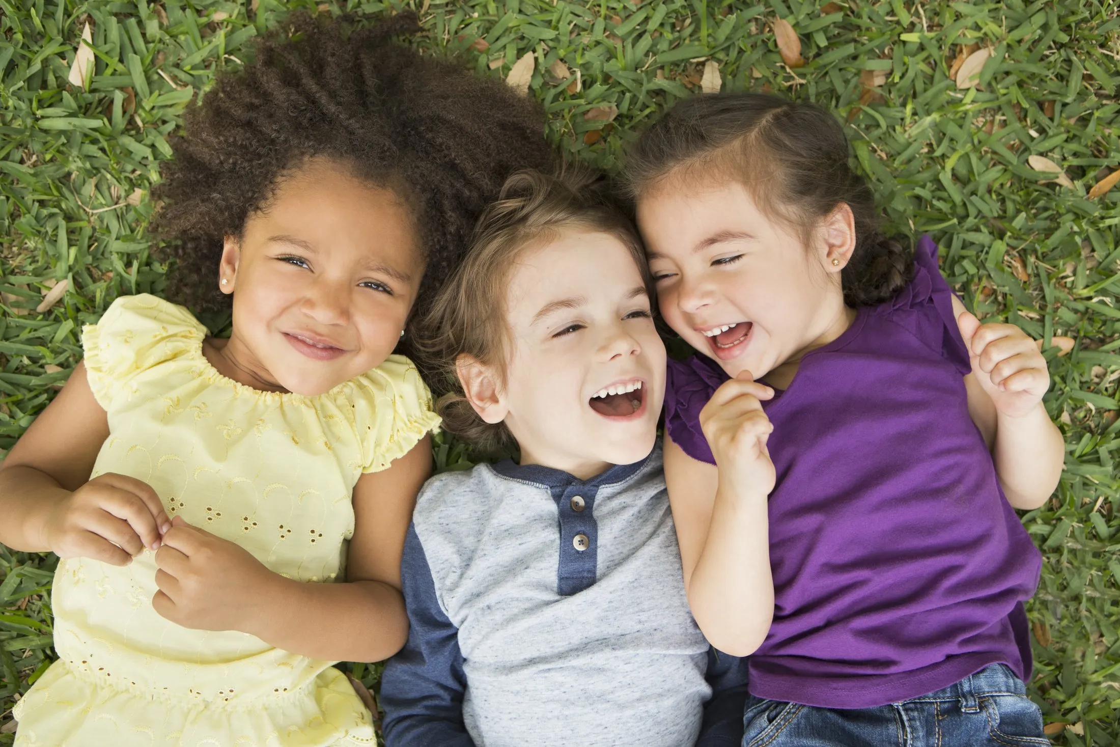 Three children lying on grass, smiling and laughing, wearing yellow, gray, and purple shirts.