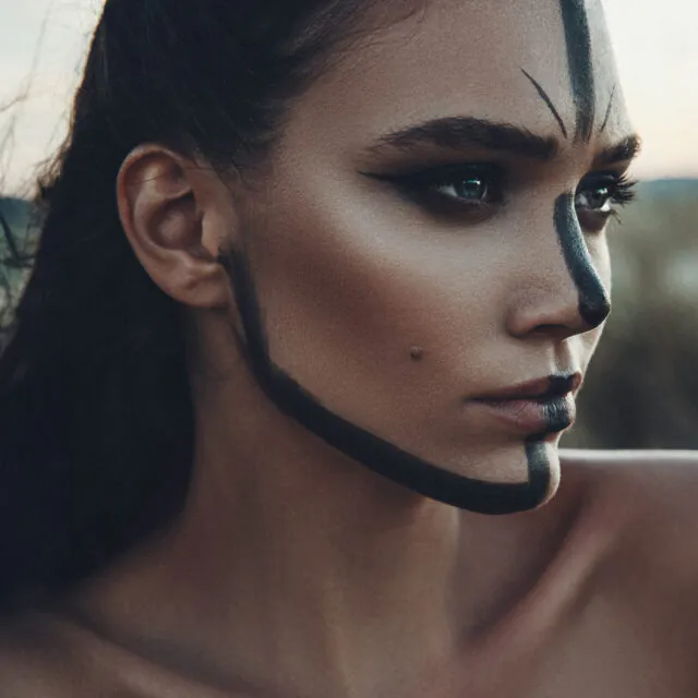 Nahaufnahme eines Frauenporträts mit schwarzer Gesichtsbemalung, die eine vertikale Linie über die Stirn und Nase sowie eine Linie entlang des Kiefers zeigt.