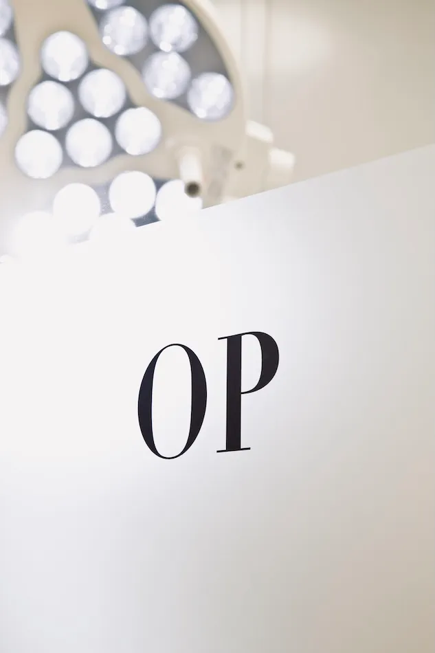 Teilansicht eines OP-Raums mit leuchtendem Operationslampen im Hintergrund und der Beschriftung 'OP' an der Wand.