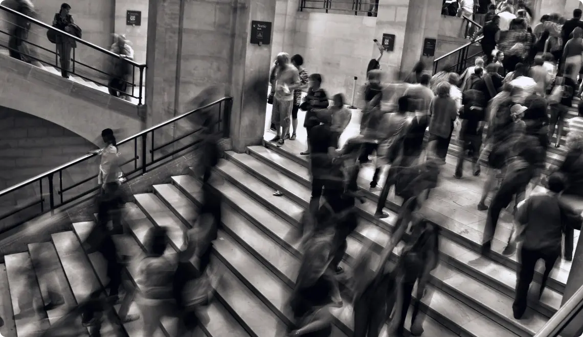 Blurred black-and-white image of many people walking up and down wide stone stairs indoors.