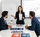 Businesswoman presenting charts to two men seated at a conference table with documents and laptop.