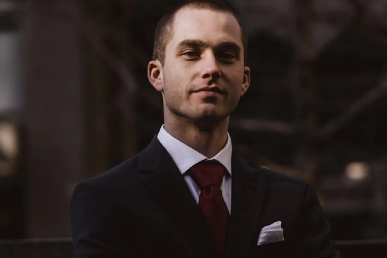 Confident man in a dark suit with a red tie and white pocket square in a blurred outdoor setting.