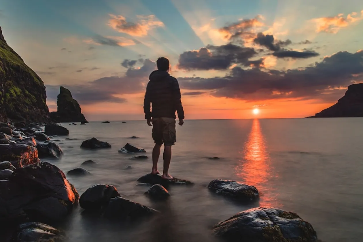 Person standing on rocks by the sea watching the sunset with rays and clouds in the colorful sky.