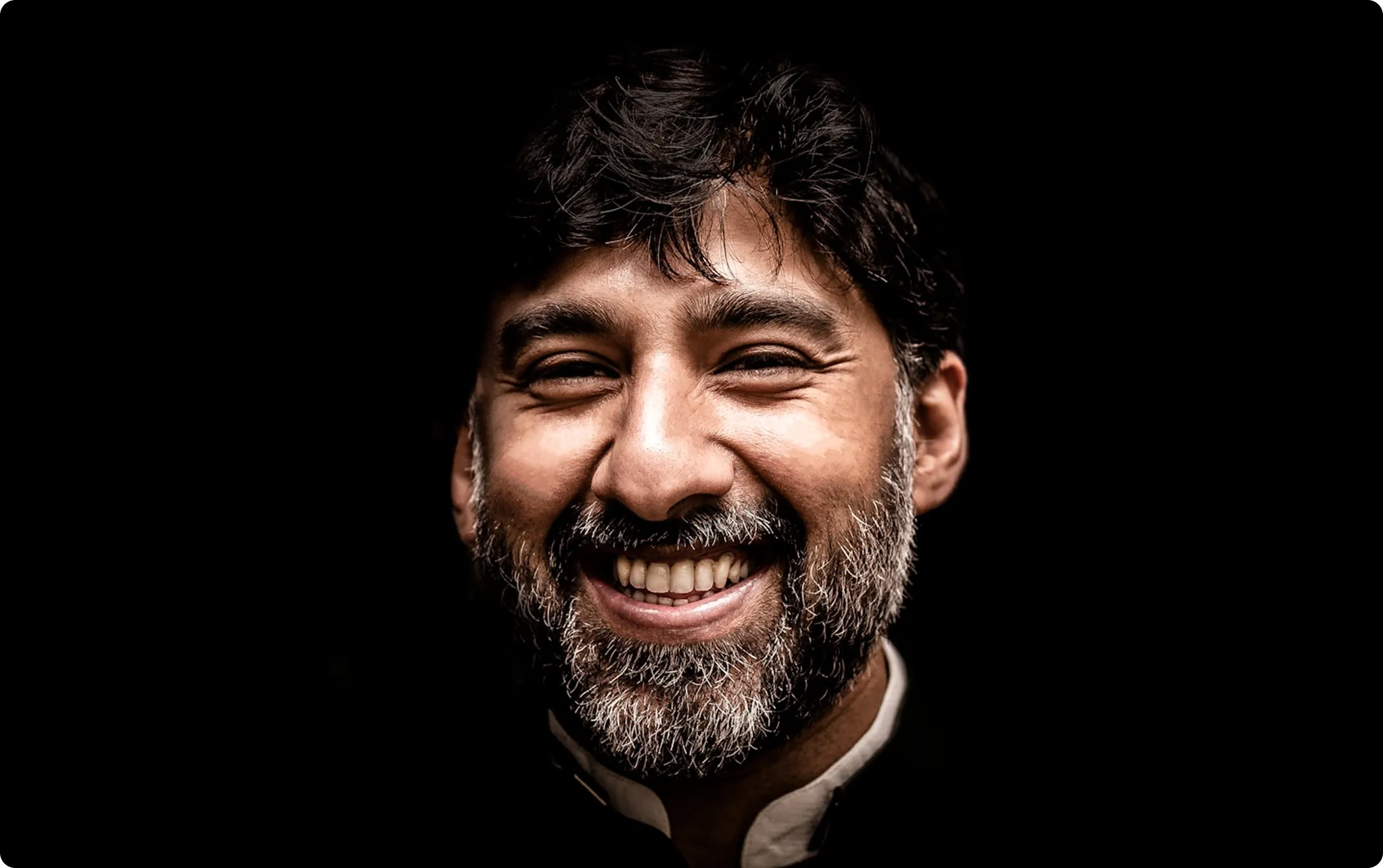 Close-up of a happy man with dark hair and a salt-and-pepper beard smiling against a black background.