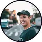 Smiling young man wearing a black cap and dark shirt outdoors in an urban setting.