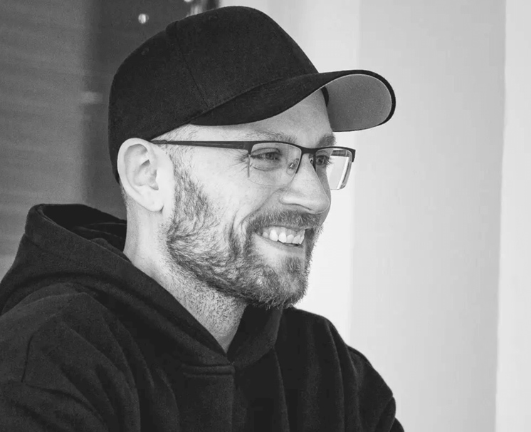 Smiling man with glasses, a beard, and a black cap wearing a black hoodie.