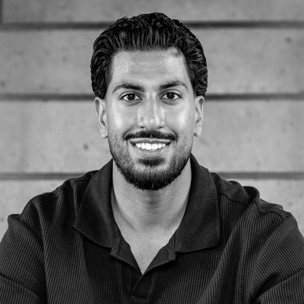 Portrait of a smiling man with short curly hair and a beard wearing a dark collared shirt against a blurred wall background.