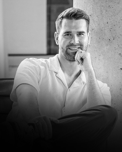Smiling man with short hair and beard leaning against a concrete pillar, dressed in a light shirt and dark pants.