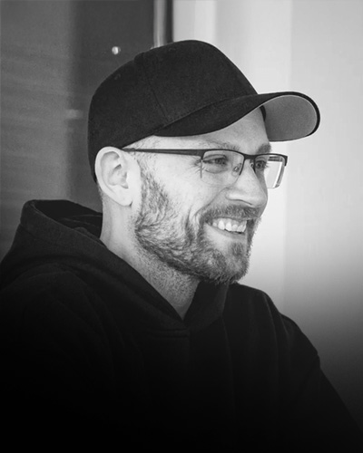 Smiling man with glasses, beard, black cap, and hoodie in a black and white photo.