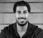 Portrait of a man with dark hair and beard, smiling, wearing a dark hoodie in front of a concrete wall.