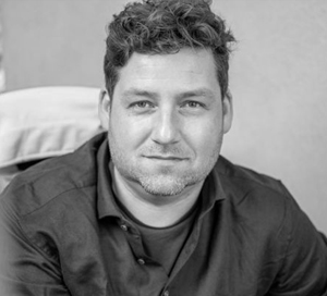 Black and white portrait of a man with short curly hair and a light beard wearing a dark shirt.