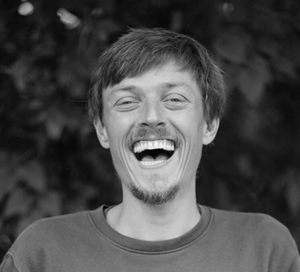 Black and white photo of a young man with short hair and a goatee laughing joyfully with blurred foliage background.