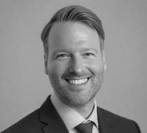 Black and white headshot of a smiling man with short hair wearing a suit and tie against a plain background.