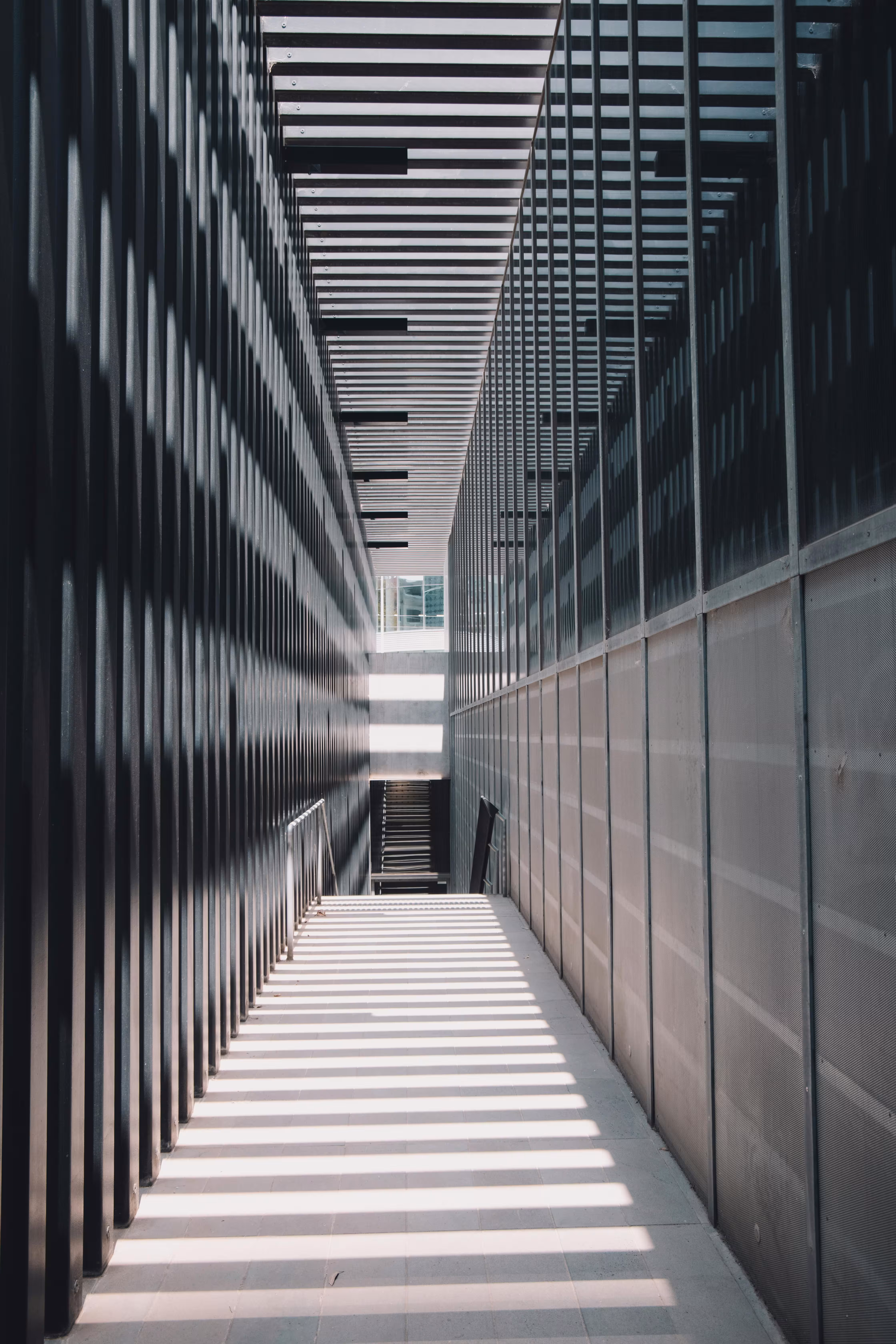 Modern corridor with metal railings and walls casting striped shadows from overhead slats.