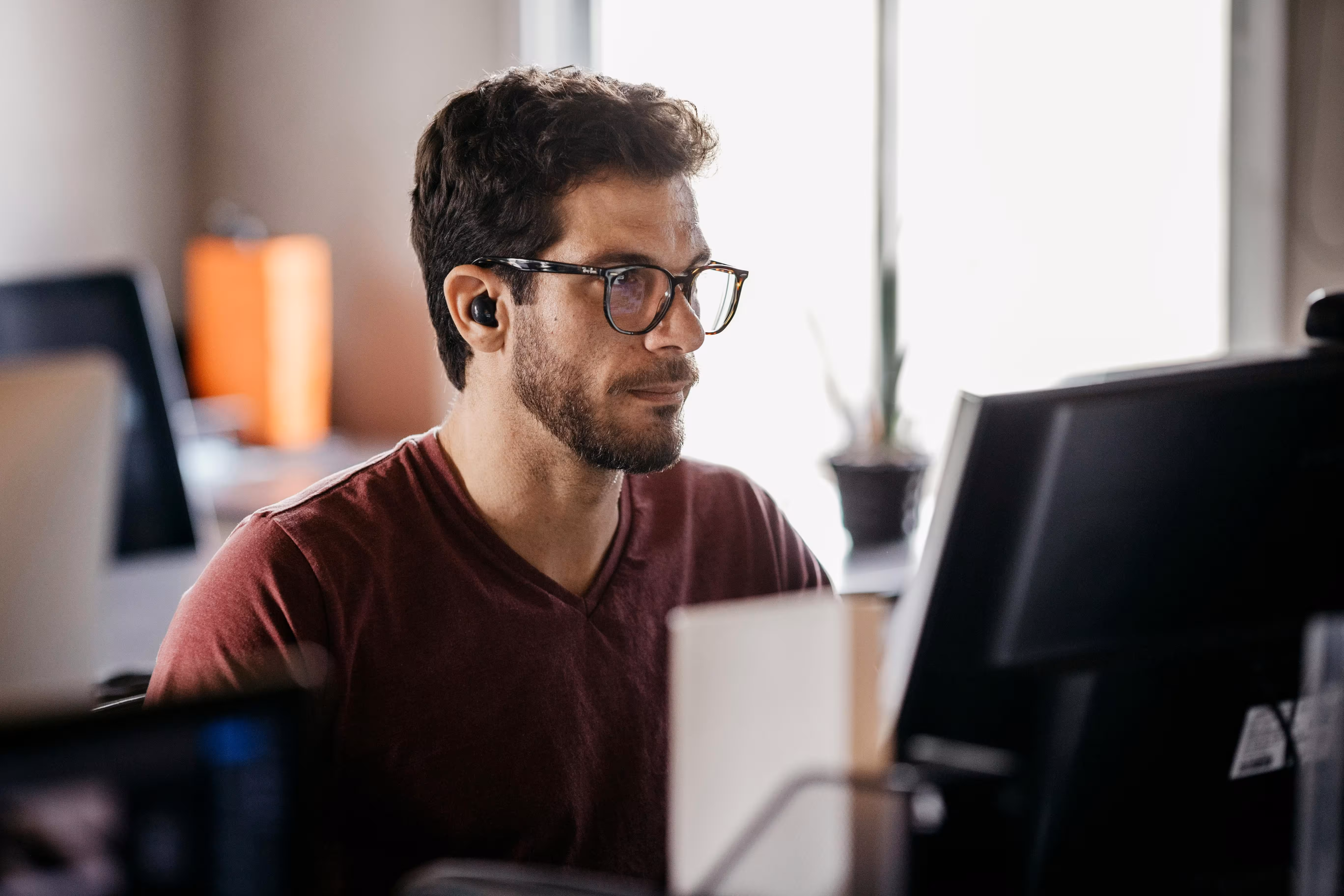 Man wearing glasses and wireless earbuds working at a computer in a modern office.