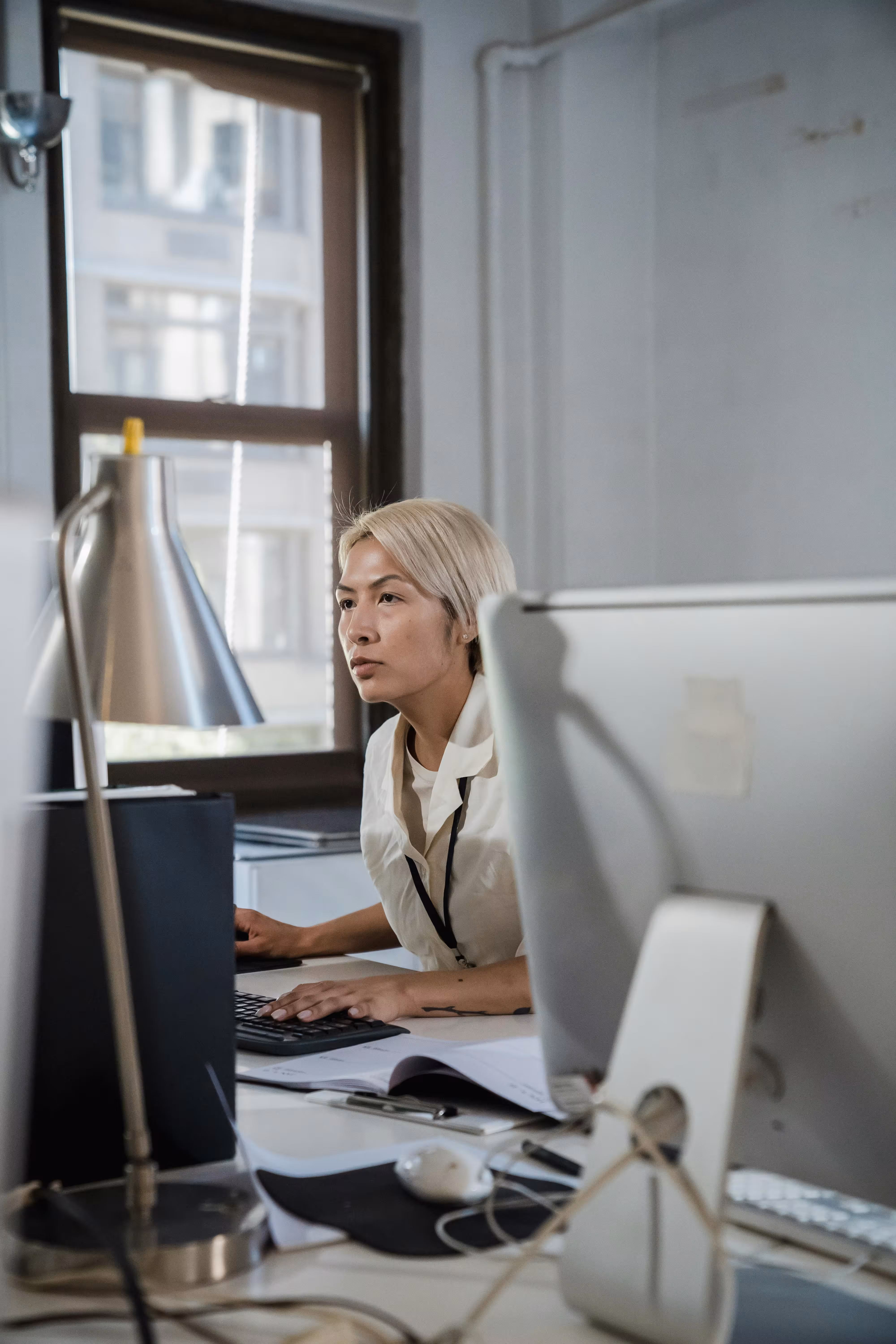 Person with short blonde hair focused on working at a computer in an office with a window in the background.