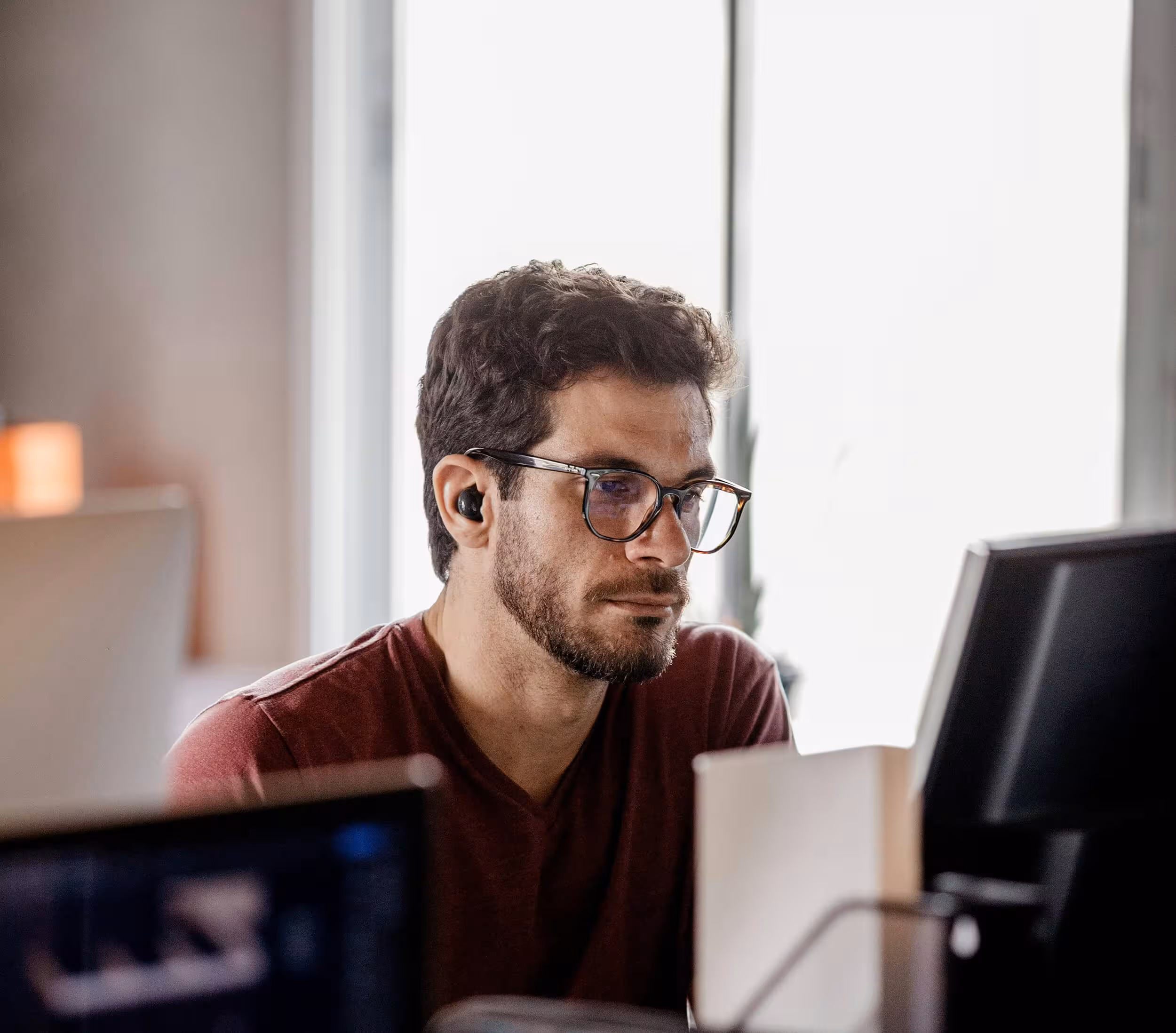 Man with glasses and earbuds focused on a computer screen in a softly lit room.