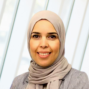 Smiling woman wearing a gray blazer in front of a light background with vertical panels.