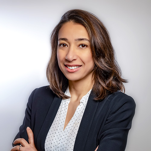 Woman with shoulder-length brown hair wearing a black blazer and white polka dot blouse, smiling with arms crossed.