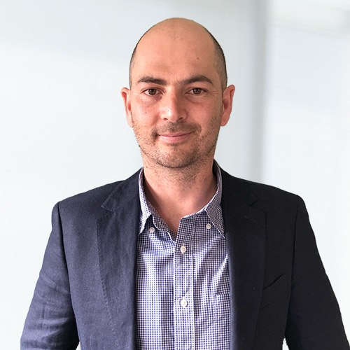 Smiling man wearing a navy blazer over a checkered shirt against a light background.