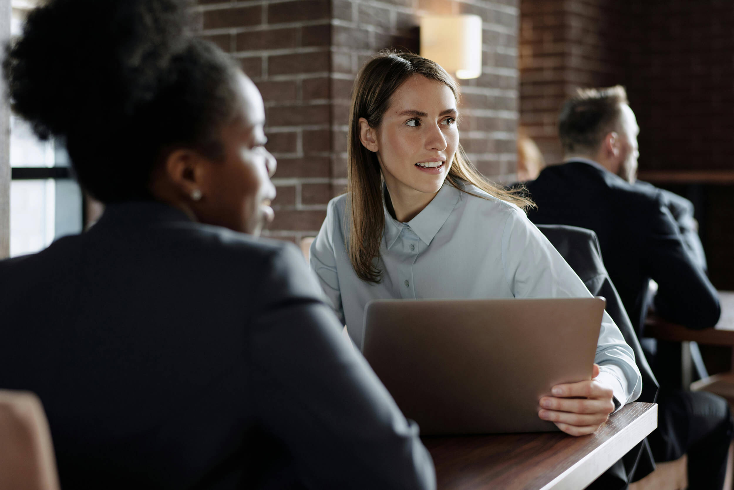 Two professional women having a discussion in an office setting, with one holding a laptop and a man sitting in the background.