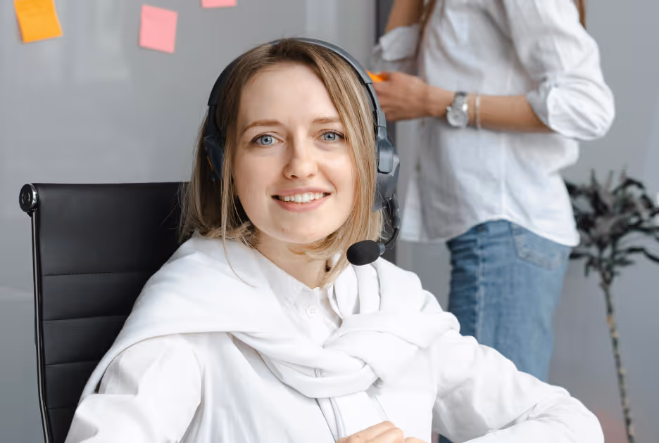 Smiling woman with headset sitting in office chair, another person in white shirt and jeans in the background.