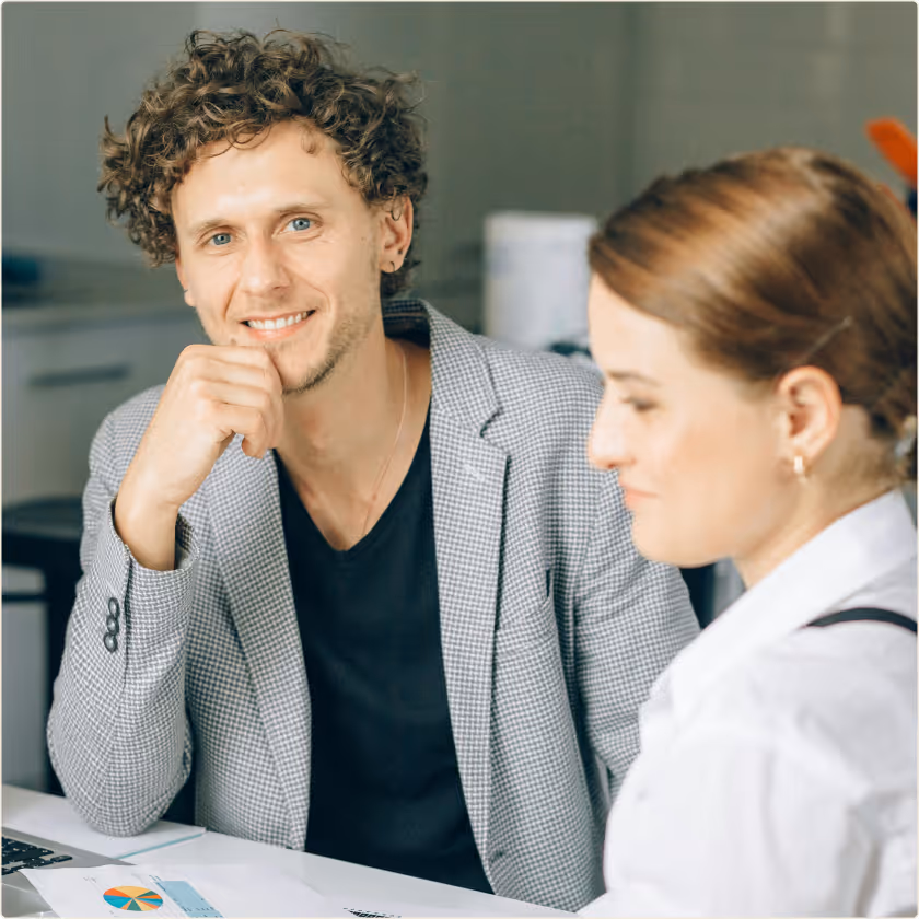 Smiling man in checked blazer sitting at a desk with a woman in a white shirt, papers and laptop nearby.