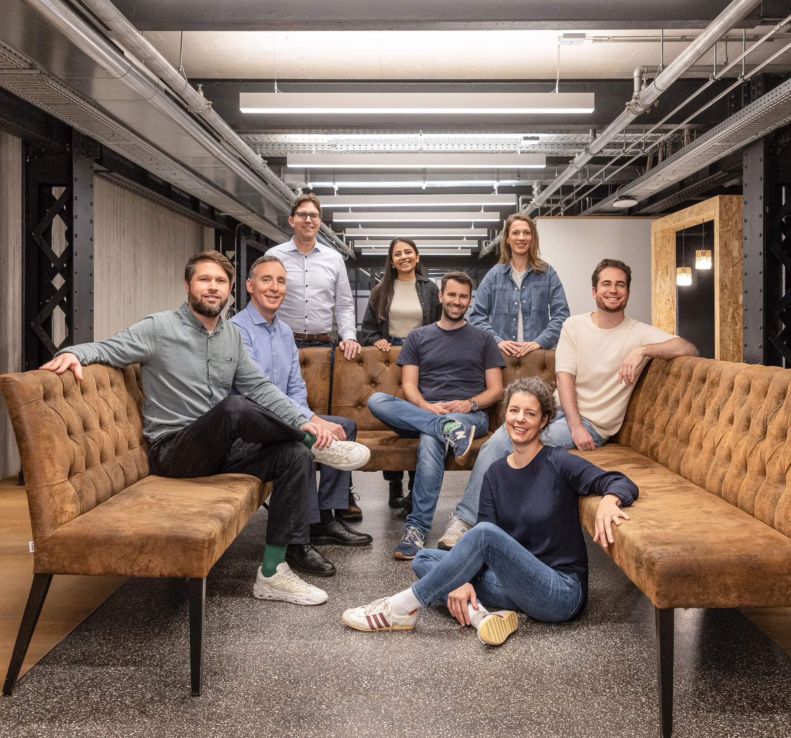 Group of eight adults smiling and posing together in a modern office lounge area with brown sofas and overhead lighting.