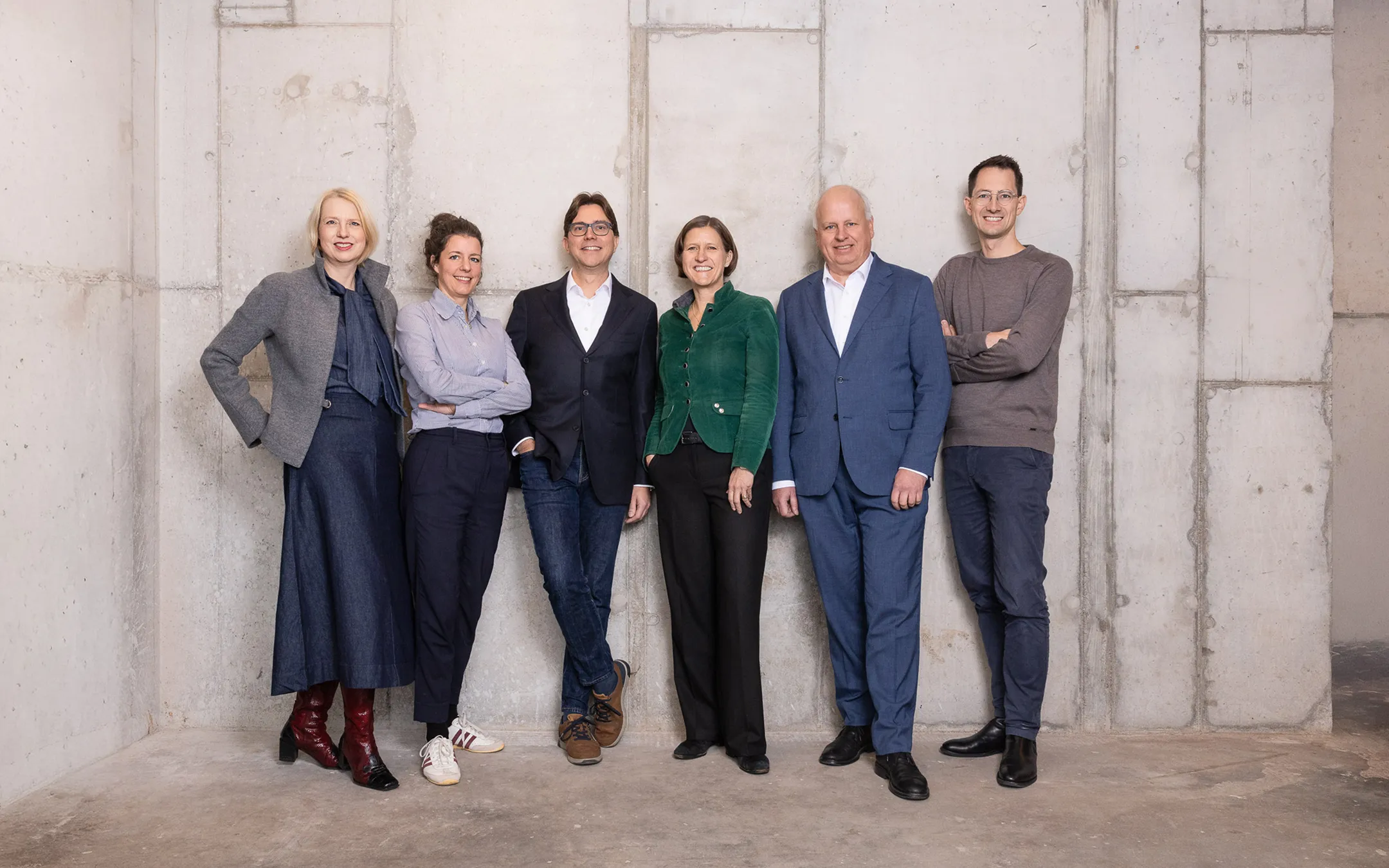 Group of six professionally dressed people standing against a plain concrete wall, smiling and facing the camera.