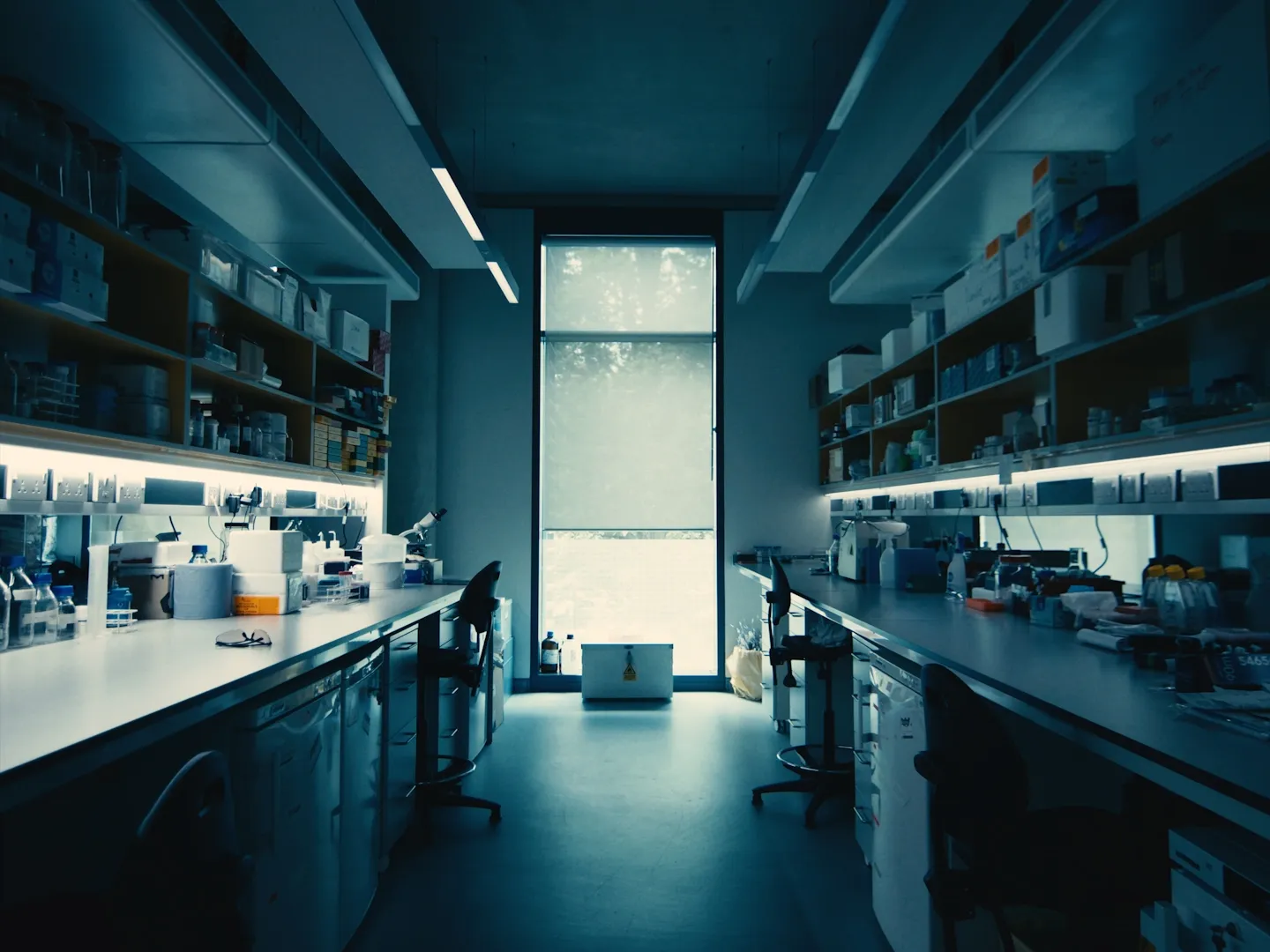 Interior of a scientific laboratory with long benches, chairs, shelves with supplies, and a tall window with a blind.