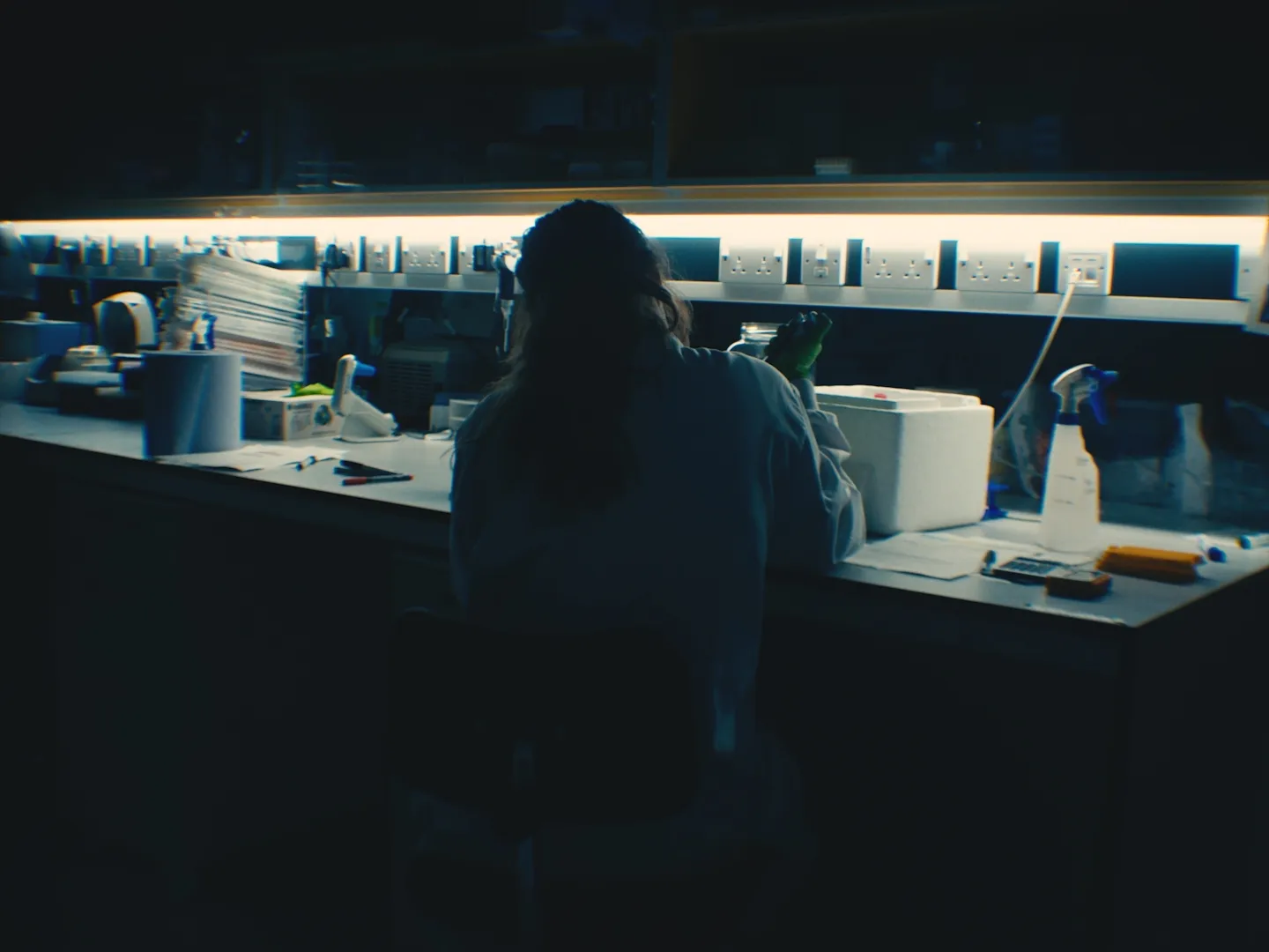 Person in a lab coat and green gloves working at a laboratory bench with equipment and supplies, illuminated by under-shelf lighting.
