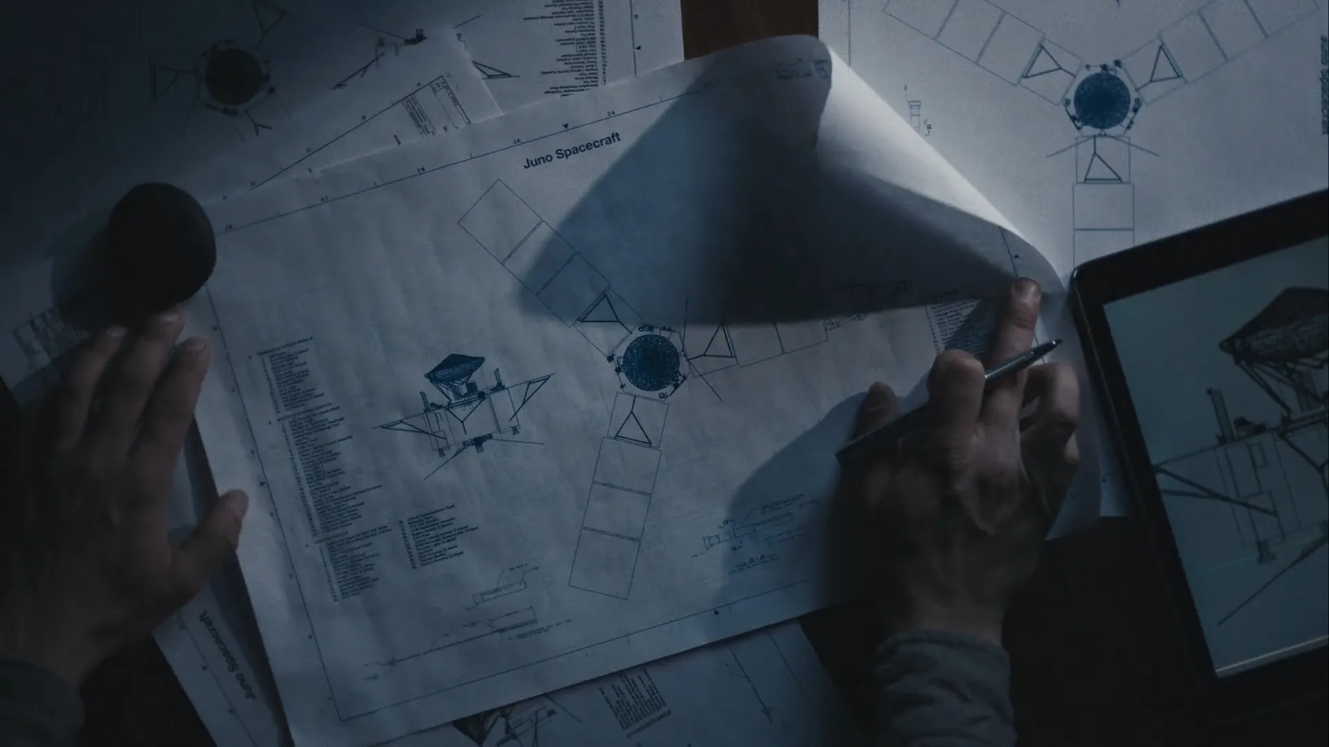 Hands holding and reviewing technical blueprints and a digital schematic of the Juno Spacecraft on a dark desk.
