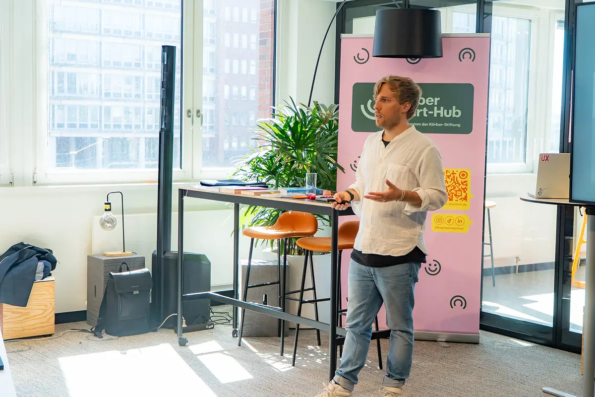 Man in casual clothes giving a presentation in a modern office with tall windows, orange stools, and a pink Körber Start-Hub banner.