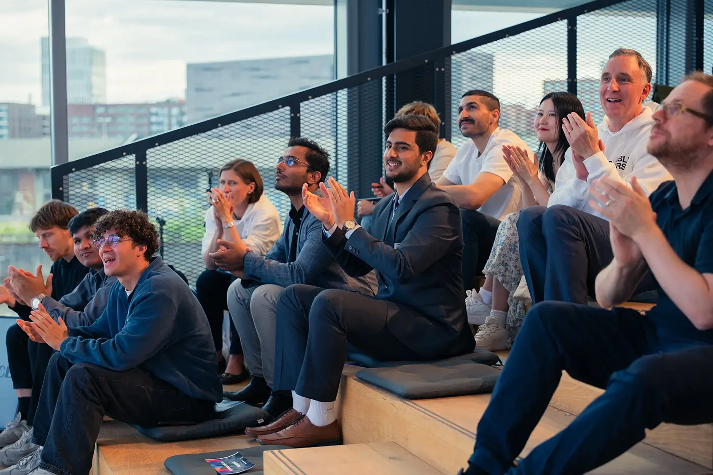 A diverse group of people seated on tiered wooden benches indoors, clapping and smiling, engaged in an event.