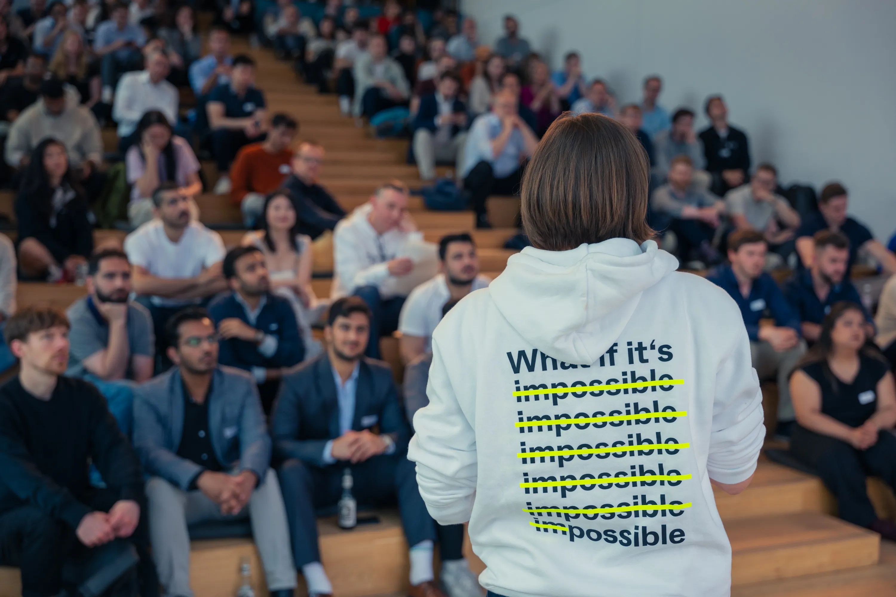 Person facing an audience wearing a white hoodie with the text 'What if it's' and the word 'impossible' repeatedly crossed out in yellow except once spelled as 'possible'.