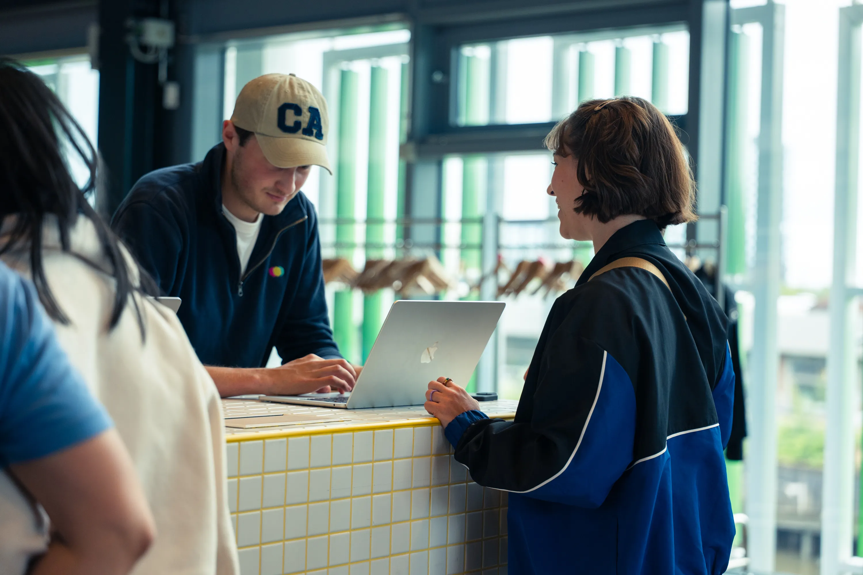 Man wearing a beige cap and navy jacket working on a laptop, talking to a woman in a blue and black jacket at a tiled counter.