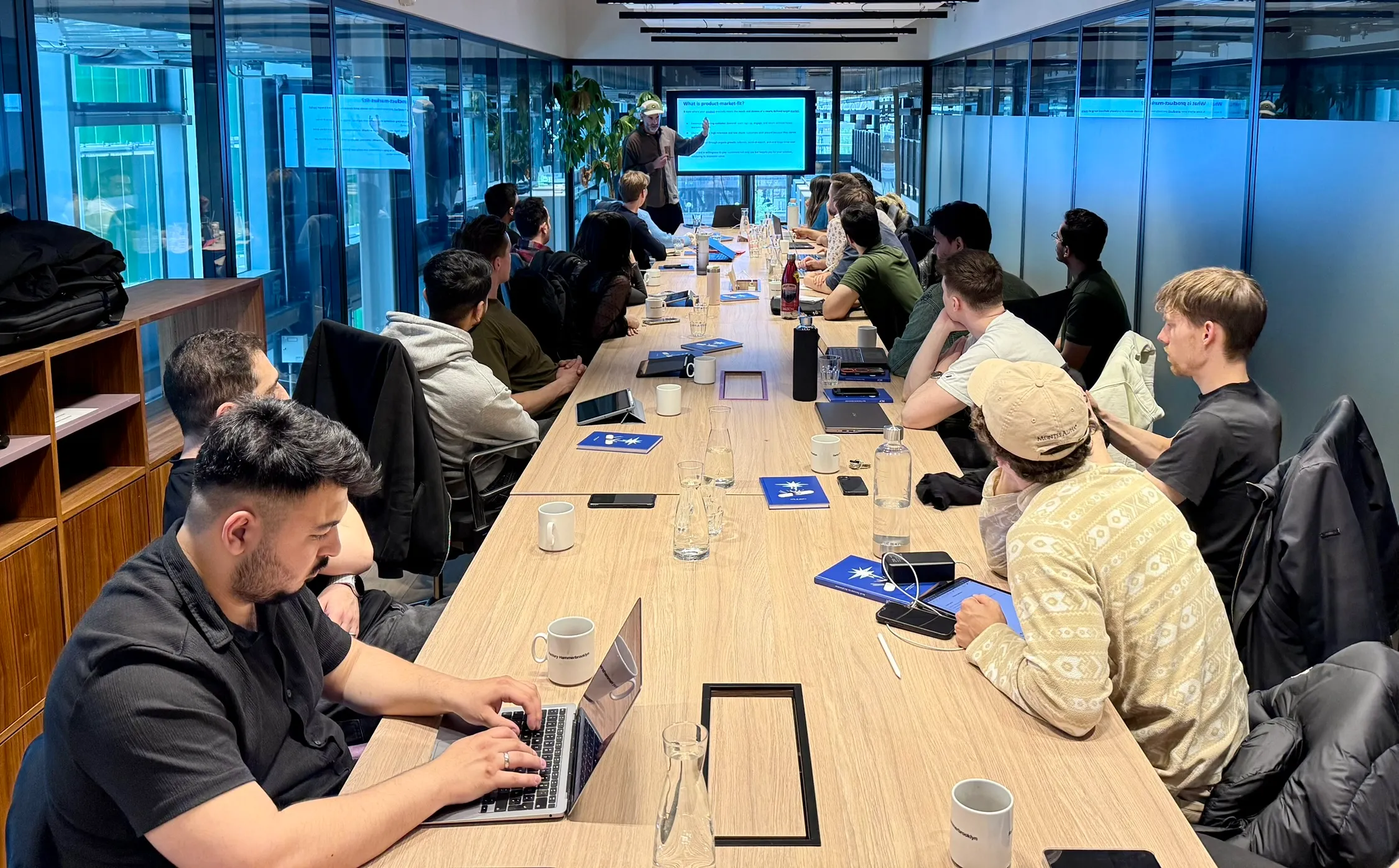 A group of diverse people seated around a long conference table attentively listening to a presenter standing near a large screen displaying a presentation.