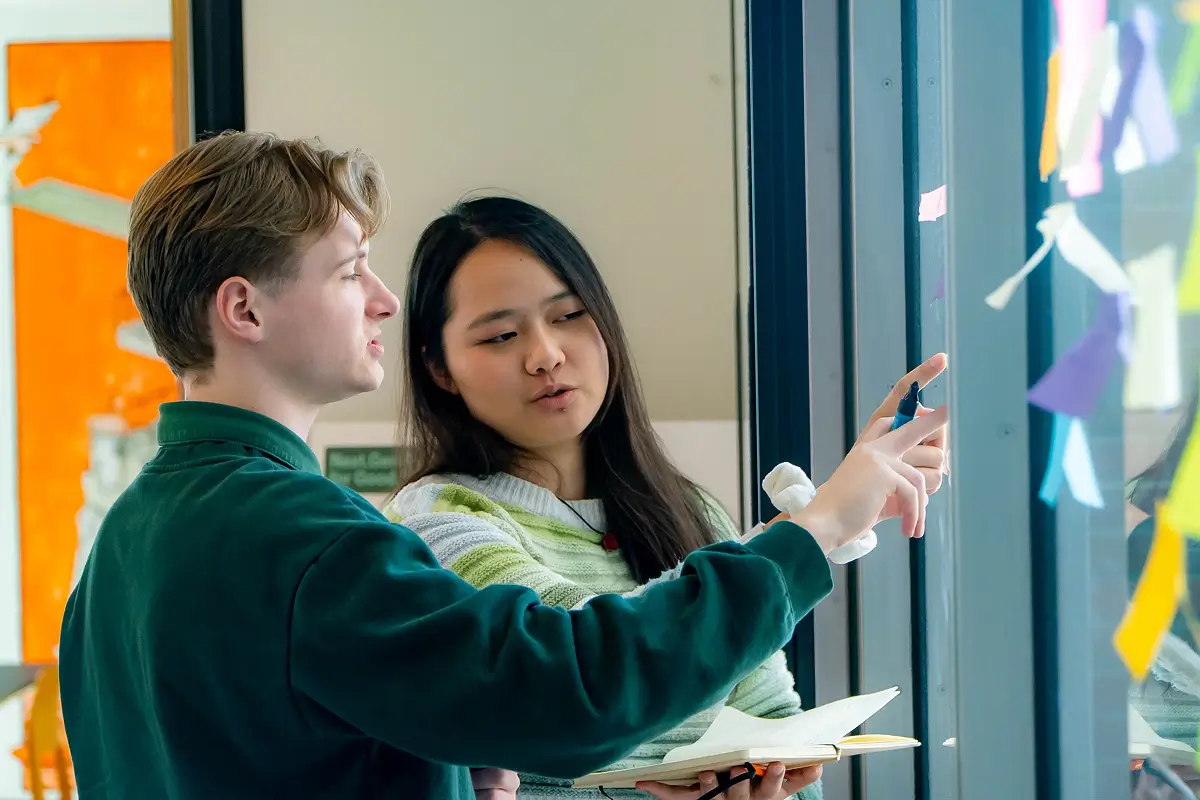 Two young people collaborating, pointing at notes on a glass wall while holding a notebook and marker.