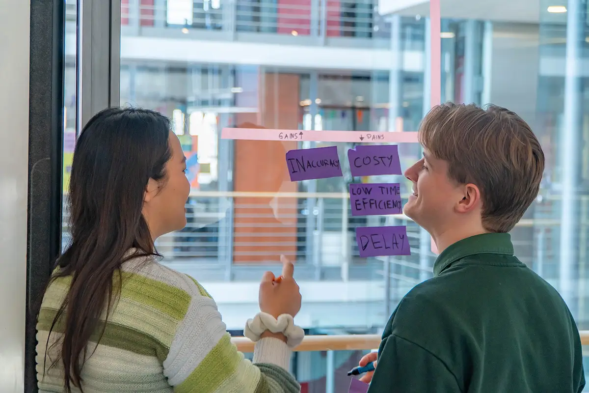 Two colleagues discussing ideas with sticky notes labeled 'Inaccuracy', 'Costly', 'Low Time Efficiency', and 'Delay' on a glass wall.