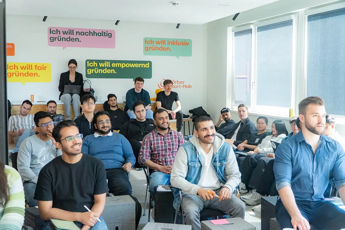 A diverse group of young adults sitting in a bright room with large windows, attentively participating in a workshop or seminar.