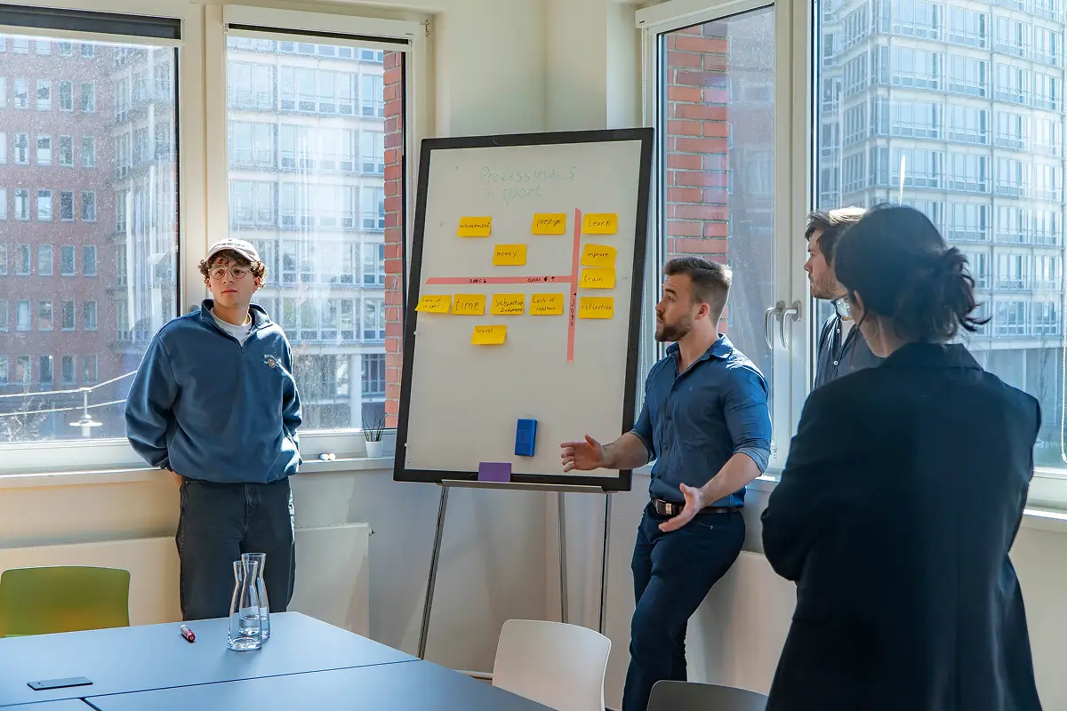 Four people having a discussion around a whiteboard with sticky notes in a bright office room with large windows showing city buildings outside.