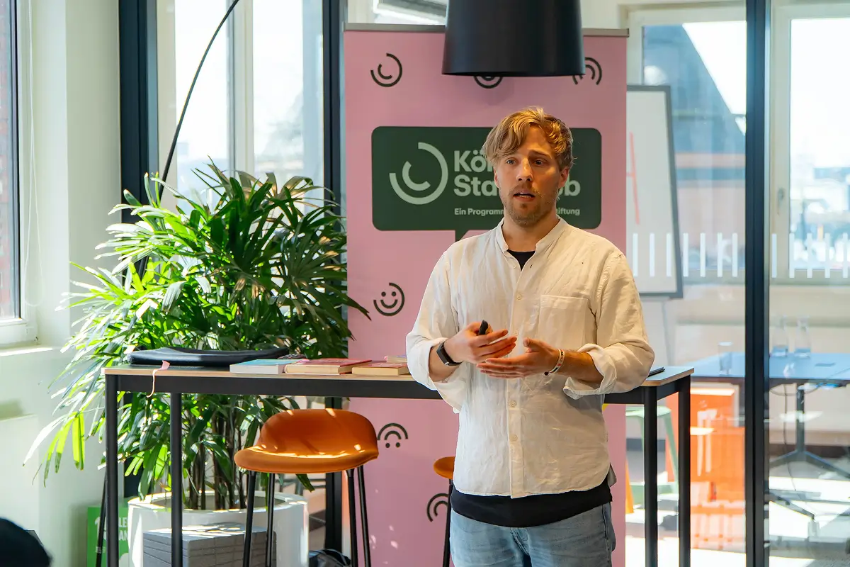 Man in a white shirt giving a presentation in a modern room with large windows and a plant.