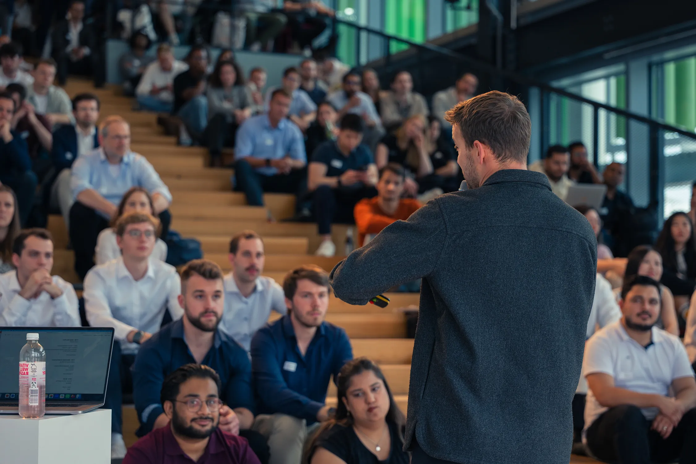 Man speaking to an audience seated on wooden steps in a modern indoor space, with a laptop and water bottle in the foreground.