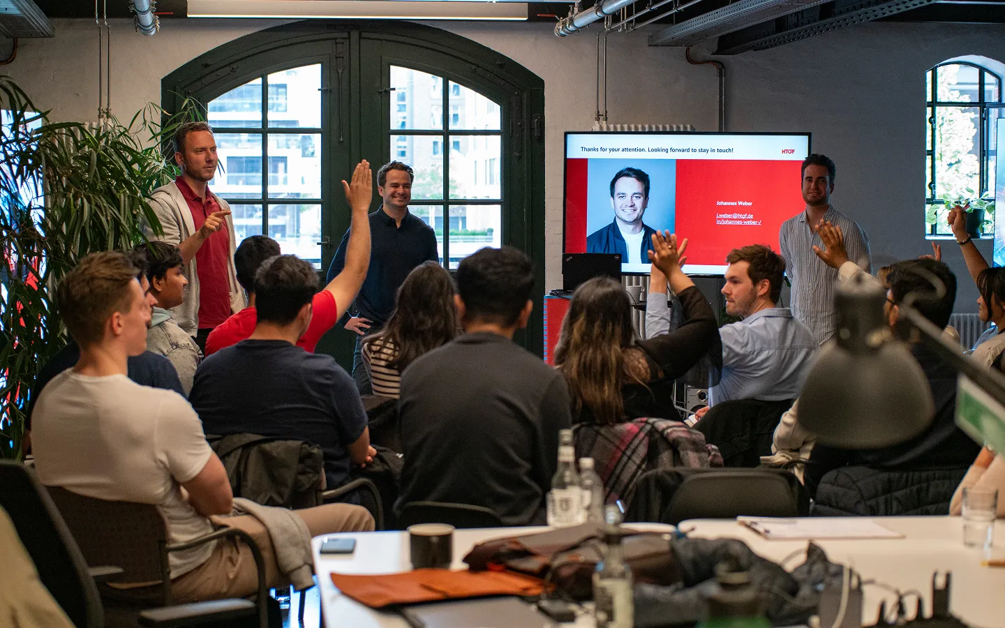 Group of people attending a presentation in a modern room with large windows, some raising hands, and a screen showing a closing slide with a man's photo and contact information.