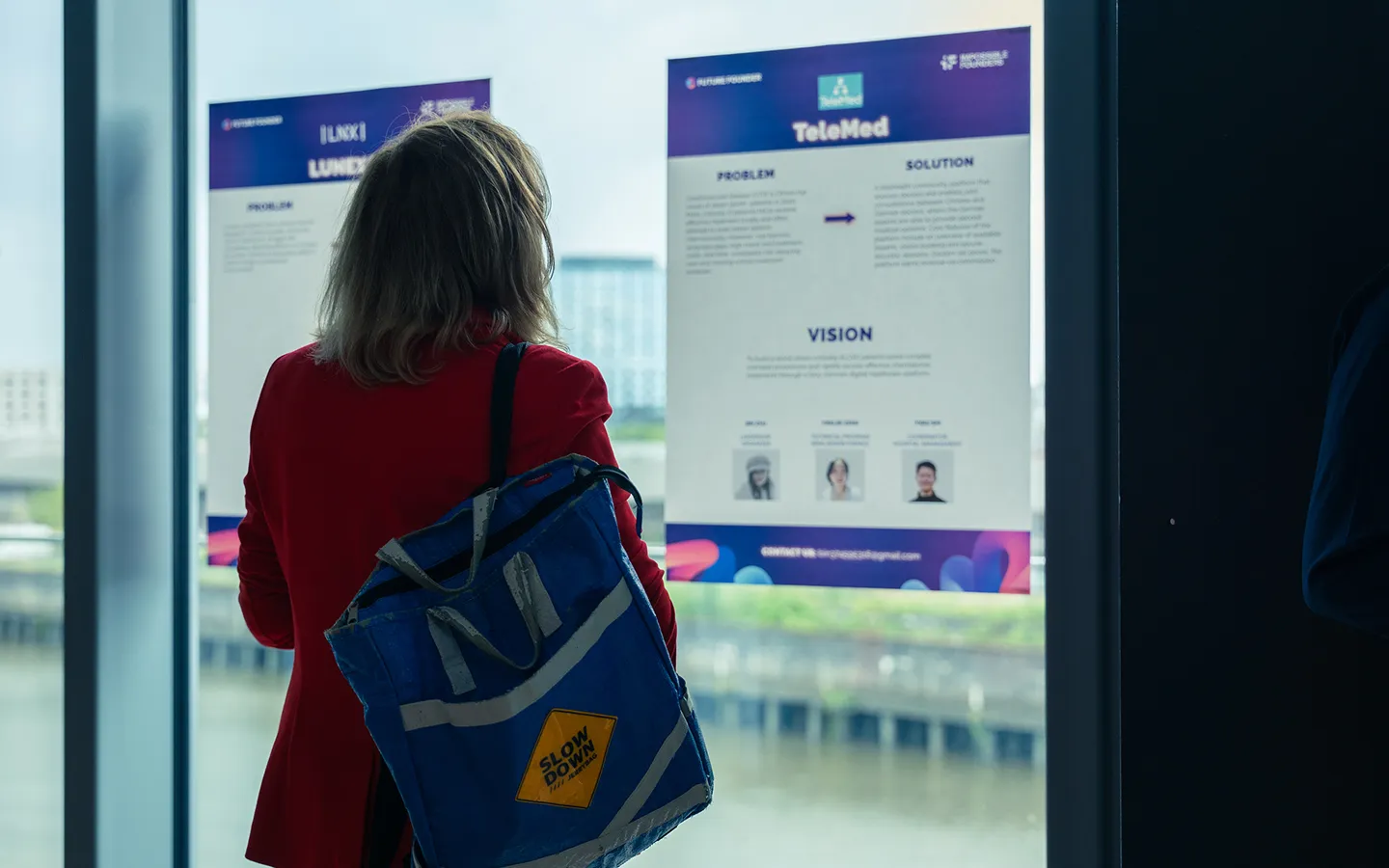 Person in red jacket with a blue bag reading informational posters about telemedicine on a window overlooking a waterfront.